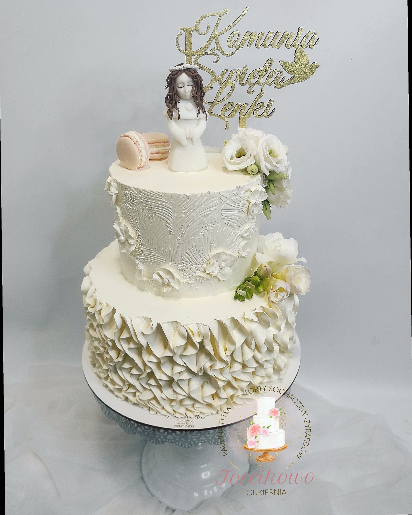 Dwupiętrowy tort komunijny w bieli, zdobiony falbanami, kwiatami i figurką dziewczynki w sukience, z topperem 'Komunia Święta Lenki' i beżowym makaronikiem