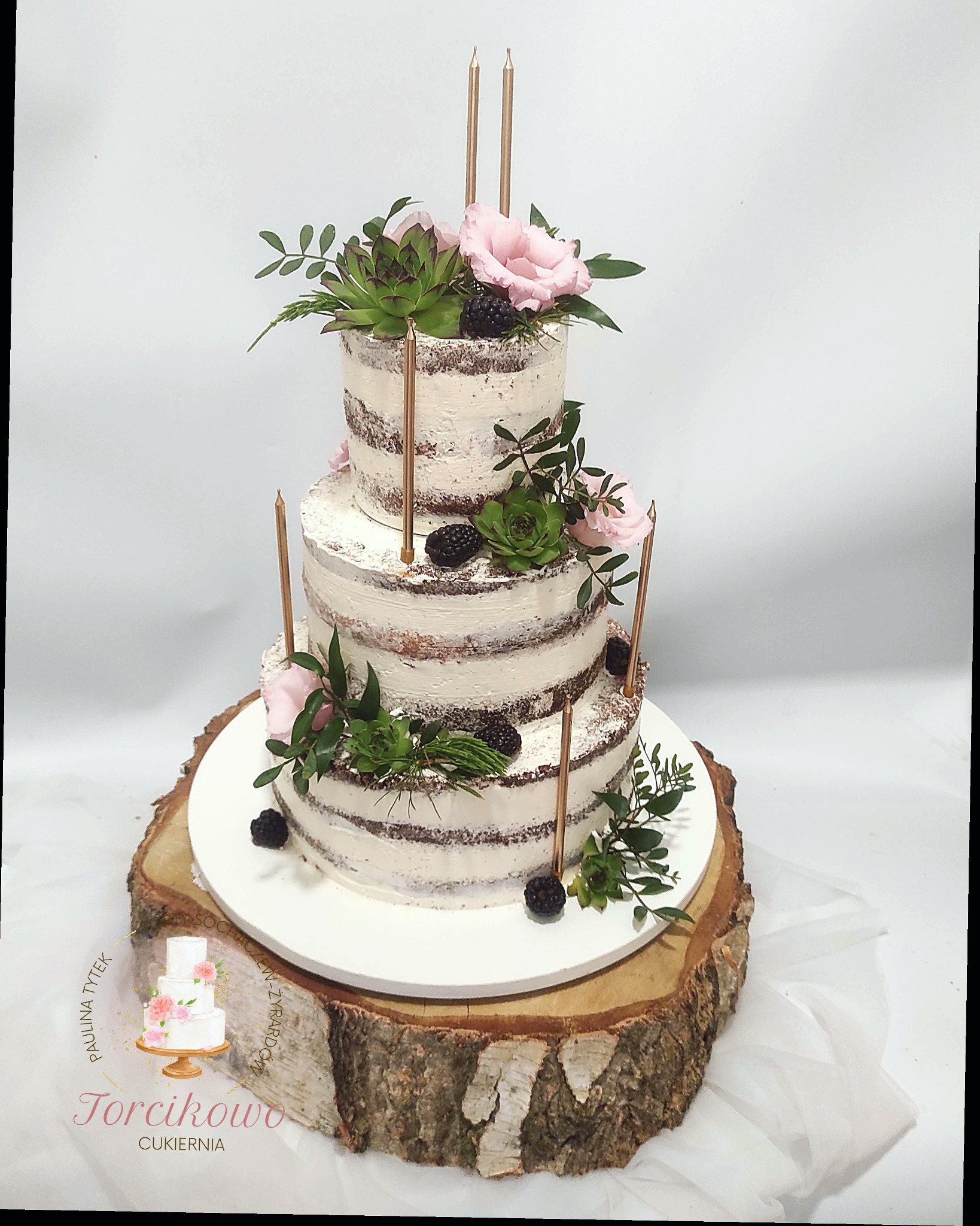 Trzypiętrowy tort 'naked cake' z kremem, dekorowany różowymi kwiatami, sukulentami, jeżynami i złotymi świeczkami, umieszczony na drewnianym plastrze pnia.