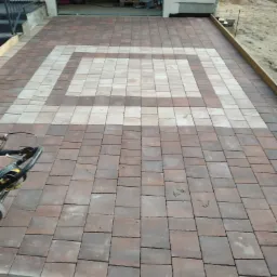 Ułożona kostka brukowa w geometryczny wzór przed garażem, widoczna ubijarka wibracyjna, przygotowane podłoże i paleta z materiałami w tle.