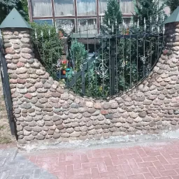 Fragment ogrodzenia z kamiennymi słupkami zwieńczonymi zielonymi daszkami, połączonych kutą, czarną bramą z ozdobnymi elementami, na tle zieleni i domu z dużymi oknami.