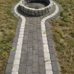 Ognisko z kamienia granitowego otoczone ścieżką z kostki brukowej w odcieniach szarości i bieli, na tle trawnika.