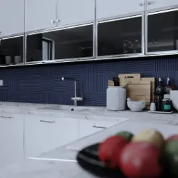 Nowoczesna kuchnia z białymi szafkami, ciemnoniebieskimi płytkami na ścianie i marmurowym blatem; na blacie widoczne akcesoria kuchenne i miska z owocami na pierwszym planie.