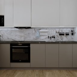 ARCHITEA. Pure Design Joanna Pura - Nowoczesna kuchnia z szarymi szafkami, marmurowym backsplash'em i drewnianą podłogą. Na blacie misa z owocami, a na ścianie zawieszone akcesoria kuchenne.