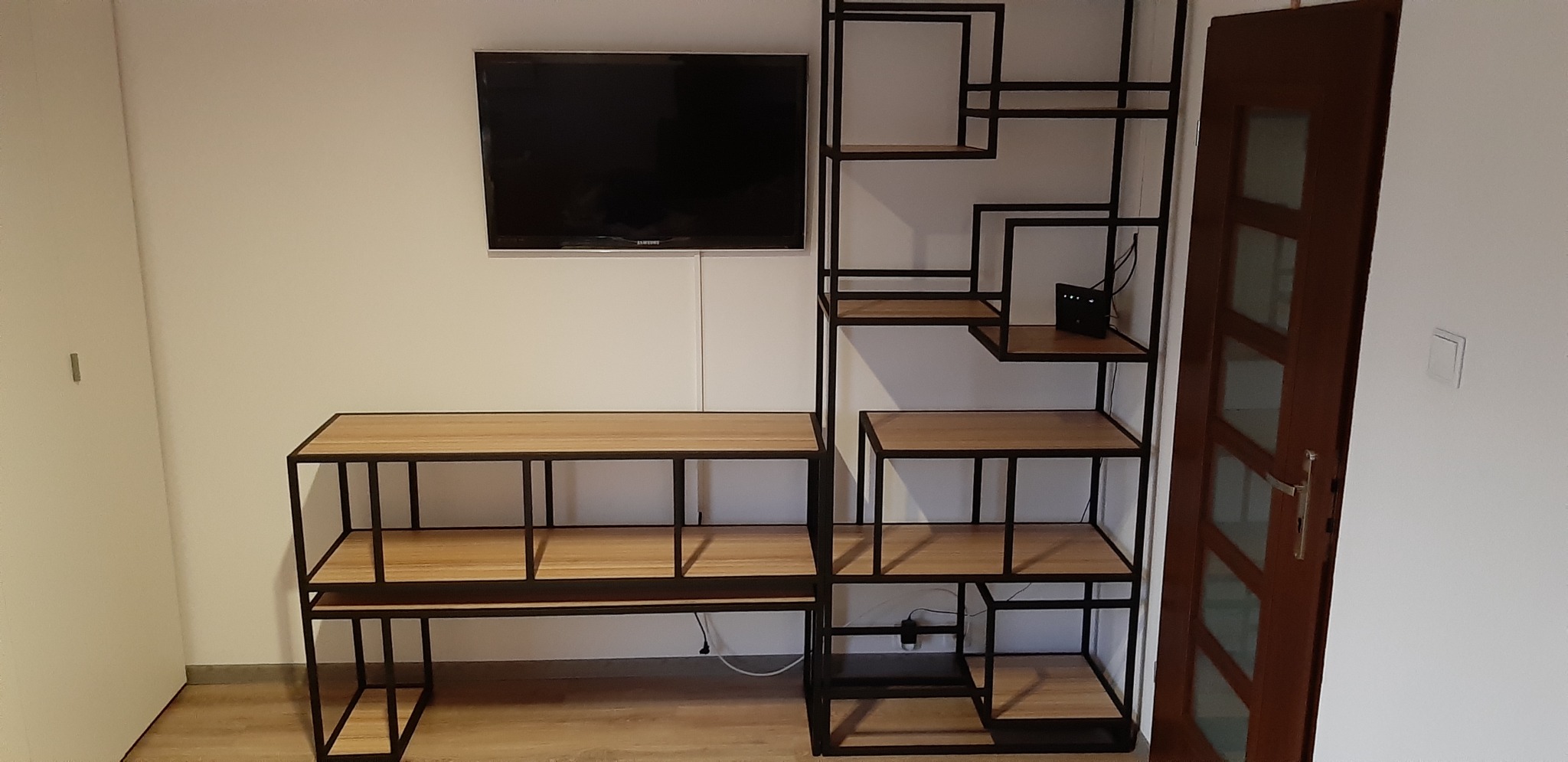 Regał z metalową ramą i drewnianymi półkami, obok szafka RTV, telewizor na ścianie i widoczny router na jednej z półek.