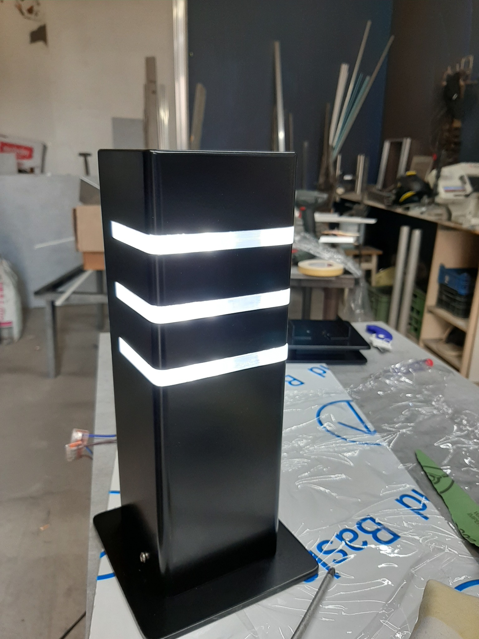 Czarny, metalowy słupek oświetleniowy z trzema poziomymi pasami światła, stojący na stole w warsztacie.