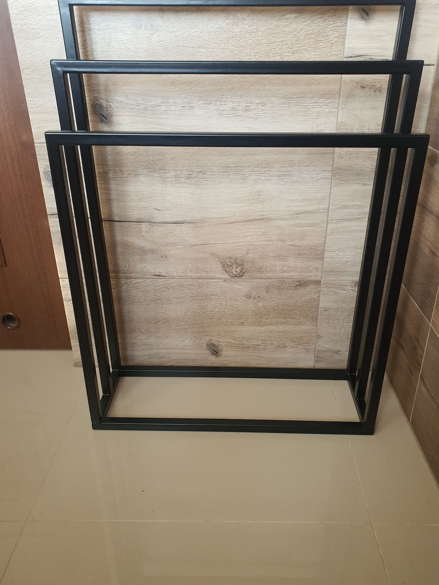 Stojak na ręczniki z czarnego metalu, spawany, widok z góry na jasnych płytkach podłogowych i ścianie z imitacją drewna.