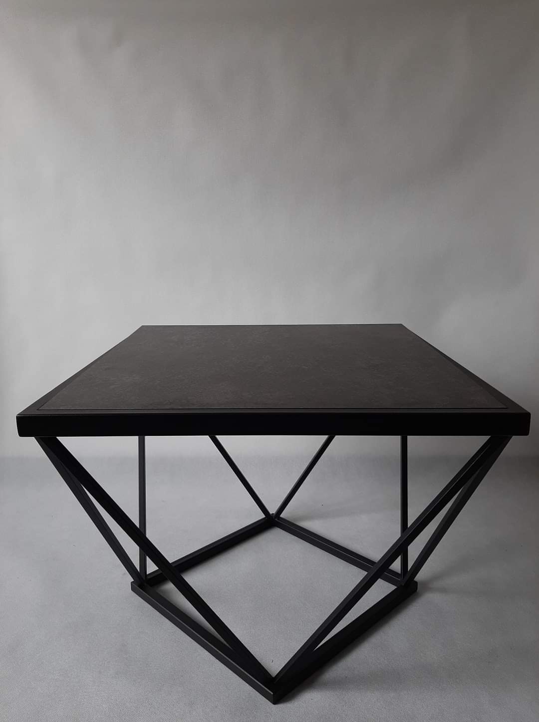 Kwadratowy stolik kawowy z ciemnym blatem i geometryczną podstawą z czarnych, metalowych prętów, ustawiony na szarym tle.