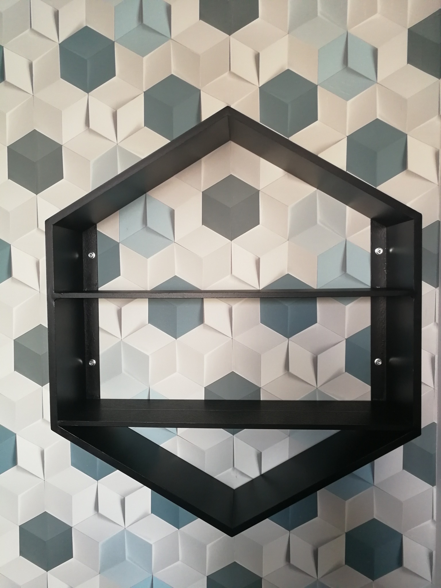 Czarna metalowa półka w kształcie domu zamontowana na ścianie z geometrycznym wzorem trójwymiarowych sześcianów w odcieniach bieli, szarości i błękitu, widoczne śruby mocujące.