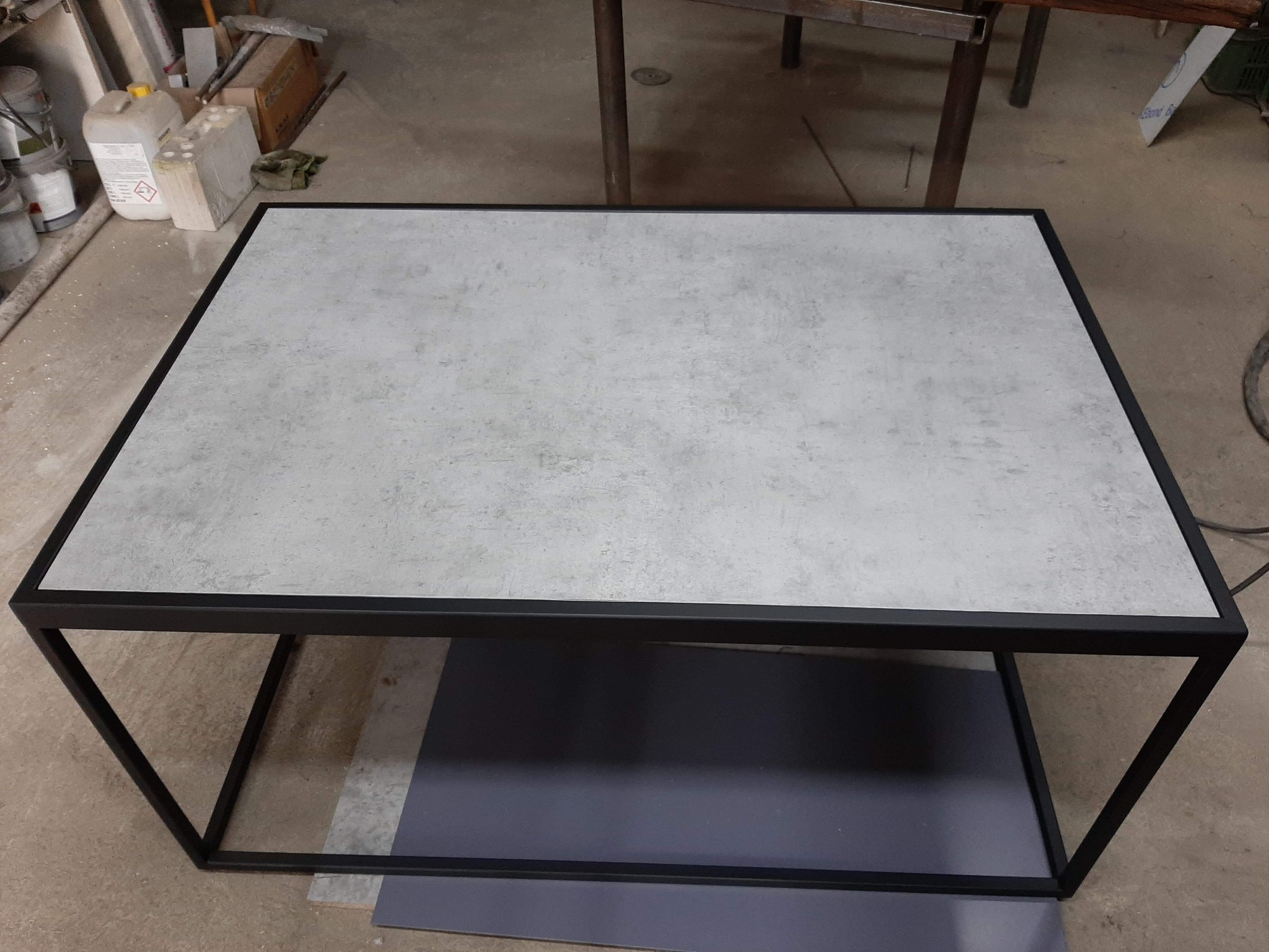 Stolik kawowy o minimalistycznym designie z blatem imitującym beton i czarną, metalową ramą, sfotografowany w warsztacie.
