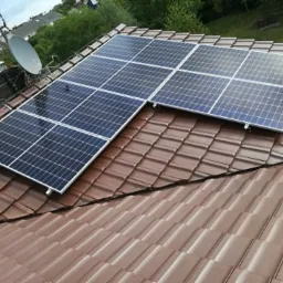 Panele słoneczne zamontowane na brązowym dachu z dachówki ceramicznej, widoczne satelita i domy w tle.