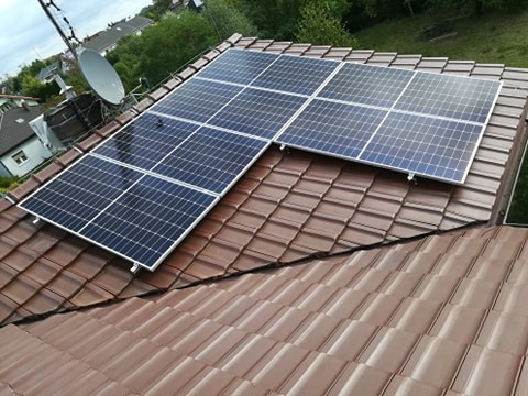 Panele słoneczne zamontowane na brązowym dachu z dachówki ceramicznej, widoczne satelita i domy w tle.