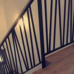 Nowoczesna, czarna balustrada schodowa z pionowymi szczeblami, zamontowana na drewnianych schodach w jasnym wnętrzu, z widocznym refleksem światła na ścianie.