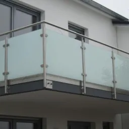 Balkon z nowoczesną balustradą ze stali nierdzewnej i matowego szkła, mocowaną za pomocą metalowych uchwytów, widok z dołu na tle białej elewacji budynku z oknami.