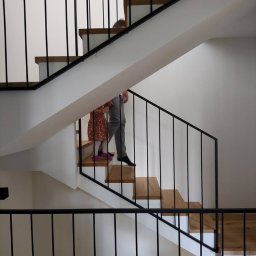 Spaw LOFT - Nowoczesne schody wewnętrzne z drewnianymi stopniami i czarnymi, pionowymi barierkami, po których schodzą dwie osoby, w minimalistycznym wnętrzu w Łomiankach.