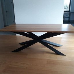 Spaw LOFT - Stół z prostokątnym, jasnym drewnianym blatem i unikalną, czarną, metalową podstawą o krzyżowym układzie nóg, na tle jasnej podłogi i białych ścian.
