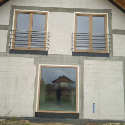 Spaw LOFT - Elewacja domu w budowie z jasnych bloczków, z dwoma oknami na piętrze z drewnianymi ramami i poziomymi, srebrnymi barierkami. W dolnym oknie odbija się osoba.