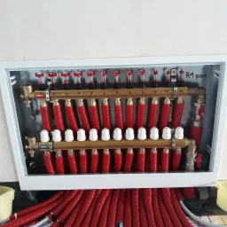 Rozdzielacz ogrzewania podłogowego w białej skrzynce z numerowanymi zaworami i rurami w czerwonej izolacji, widoczne złącza marki Ferro.