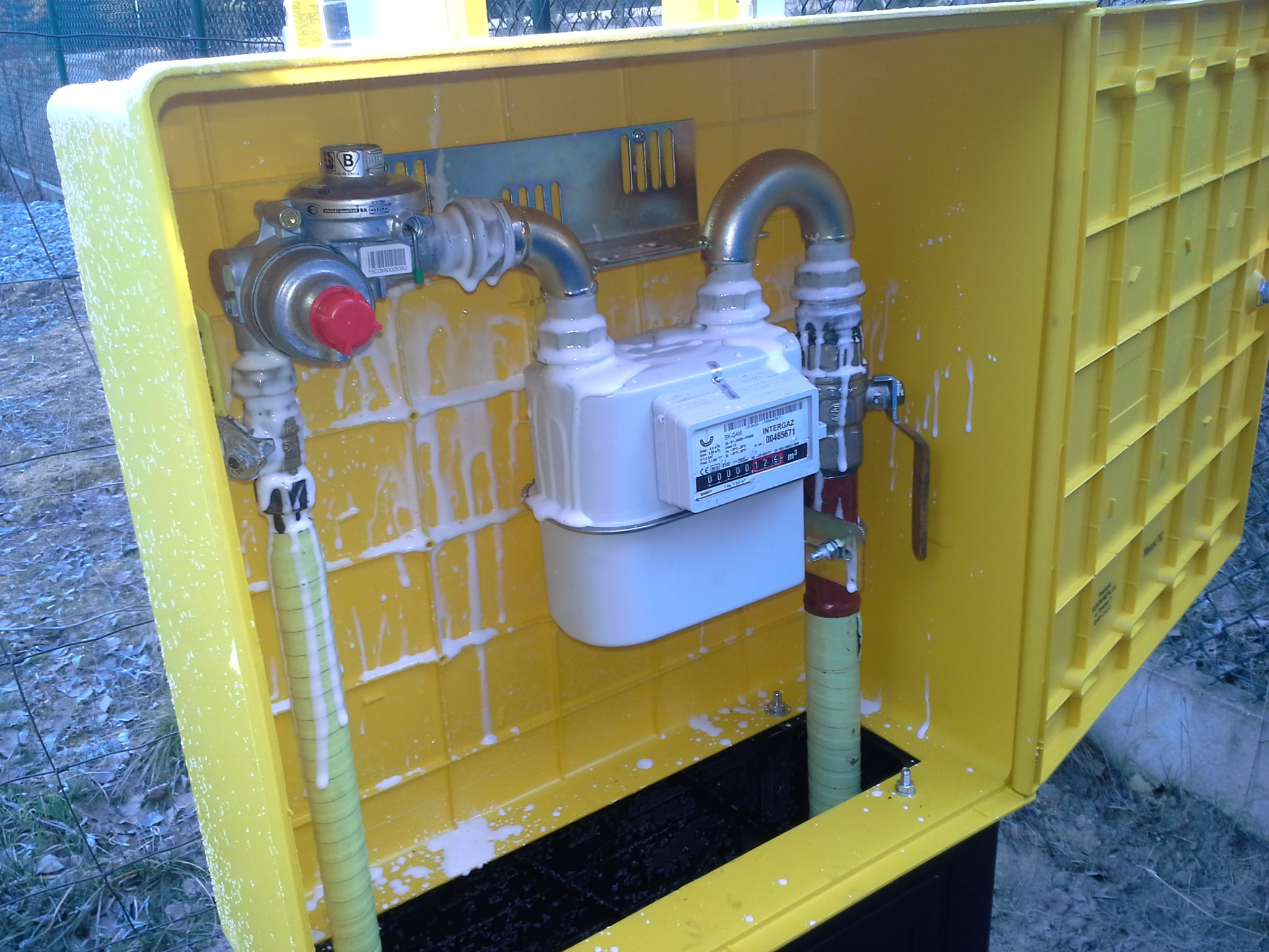 Żółta skrzynka z licznikiem gazu, rurami i reduktorem, pokryta pianą, prawdopodobnie po teście szczelności.
