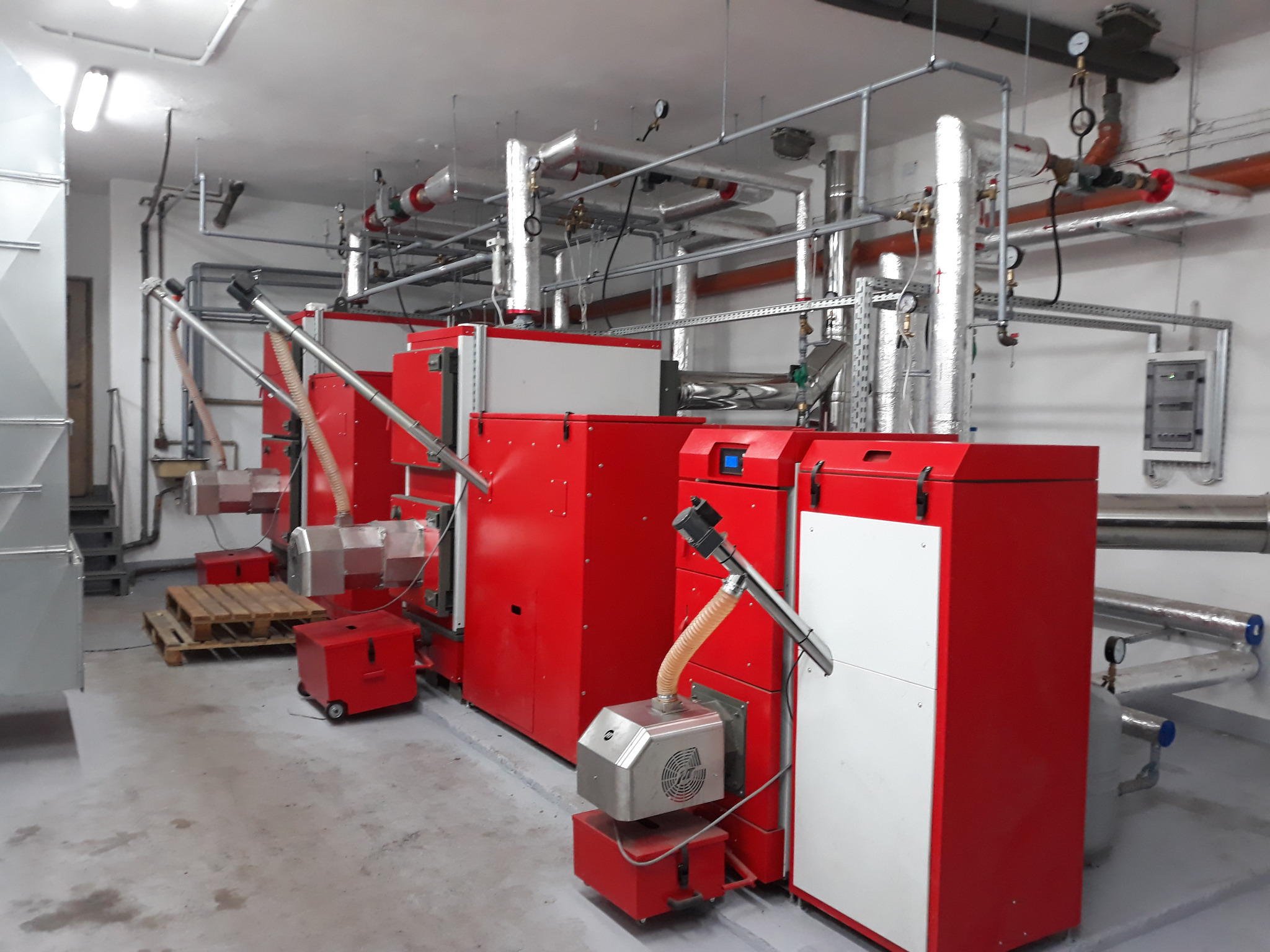 Kompleksowa kotłownia z szeregiem czerwonych kotłów na paliwo stałe, z widocznymi podajnikami, rurami instalacji grzewczej i armaturą pomiarową.