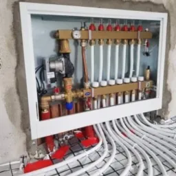 Rozdzielacz ogrzewania podłogowego z widoczną pompą obiegową, manometrem i licznymi zaworami, zamontowany w białej szafce w surowej ścianie, z licznymi białymi rurami wychodzącymi na ułożoną siatkę...