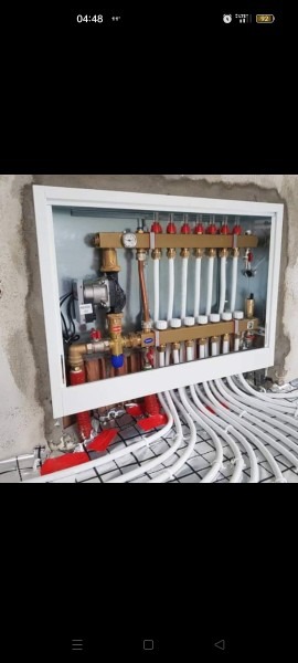 Rozdzielacz ogrzewania podłogowego z widoczną pompą obiegową, manometrem i licznymi zaworami, zamontowany w białej szafce w surowej ścianie, z licznymi białymi rurami wychodzącymi na ułożoną siatkę...