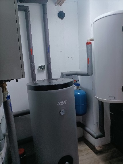 Szara pompa ciepła marki IEMEI z podłączonym niebieskim zbiornikiem wyrównawczym, widoczne rury z izolacją termiczną, instalacja w pomieszczeniu technicznym o białych ścianach.