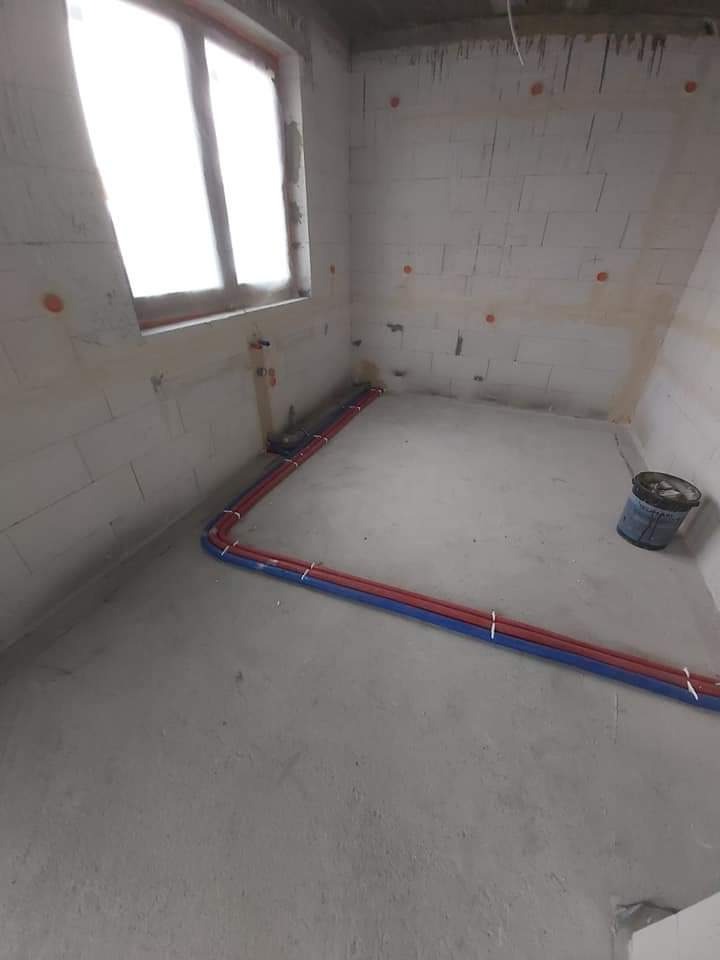 Surowy stan łazienki w trakcie prac: widoczne rury instalacji wodnej (czerwona i niebieska) prowadzone po podłodze, ściany z betonu komórkowego z zaznaczonymi punktami instalacji, okno z widokiem...