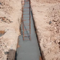 Wykop fundamentowy wypełniony świeżym betonem, z wystającą zbrojoną konstrukcją stalową, otoczony ziemią i gruzem.