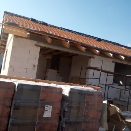 Budowa domu z białych bloczk&oacute;w: widoczne ściany, więźba dachowa z deskowaniem i częściowo ułożona dach&oacute;wka. Na pierwszym planie palety z dach&oacute;wką w folii.