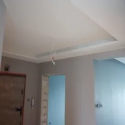 Wykończony sufit z dekoracyjnym obniżeniem i delikatnym oświetleniem, szpachlowane ściany w odcieniach szarości, widoczne przewody elektryczne przed montażem oświetlenia.