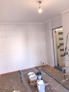 Pokój w trakcie prac: świeżo pomalowana na biało ściana, zabezpieczona folią podłoga z widocznymi plamami farby, puszki z farbą na podłodze, drabina oparta o ścianę przy drzwiach.