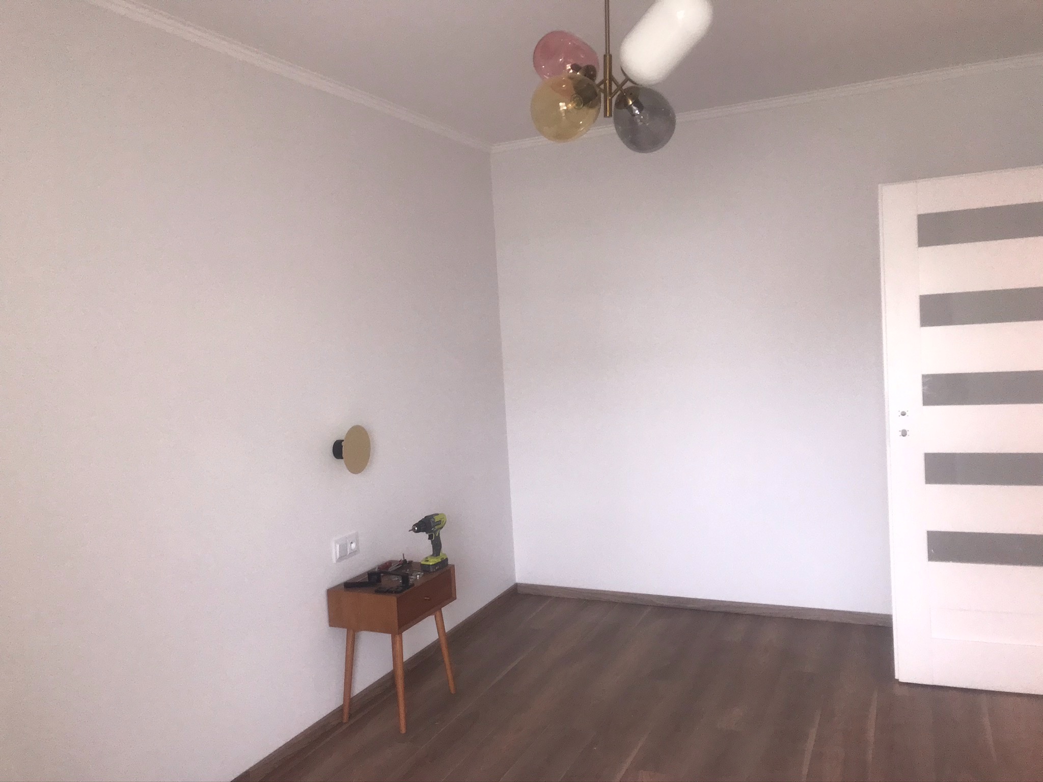 Pusta biała ściana z zamontowanym kinkietem, obok stolik z wkrętarką i narzędziami, w tle białe drzwi z poziomymi szarymi paskami i lampa sufitowa z kolorowymi kulami.