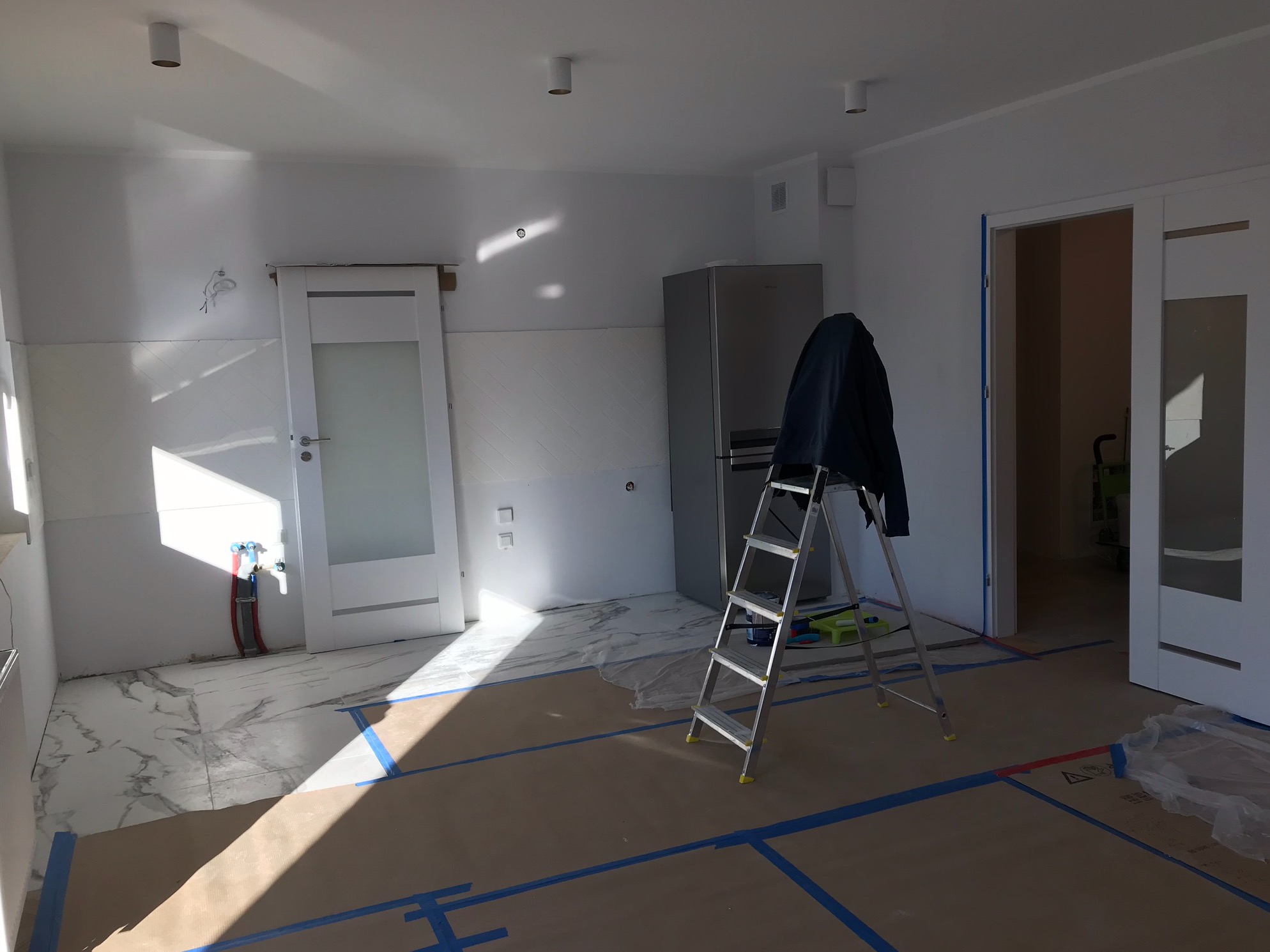 Przygotowanie do prac w jasnym pomieszczeniu: podłoga zabezpieczona kartonem i niebieską taśmą, drabina oparta o lodówkę, widoczne rury instalacji wodnej przy drzwiach.