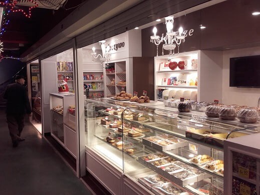 Wnętrze cukierni z przeszkloną ladą wypełnioną ciastami i deserami, podświetlane półki z produktami, ozdobne żyrandole i osoba wchodząca do lokalu.