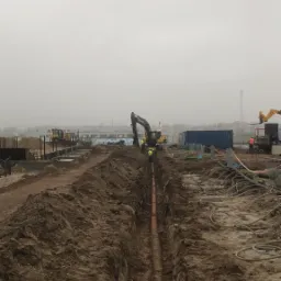 Wykonanie wykopu pod rurę drenarską, widoczny rów z ułożoną rurą, w tle pracujący sprzęt budowlany i robotnicy w odblaskowych kamizelkach, pochmurny dzień.