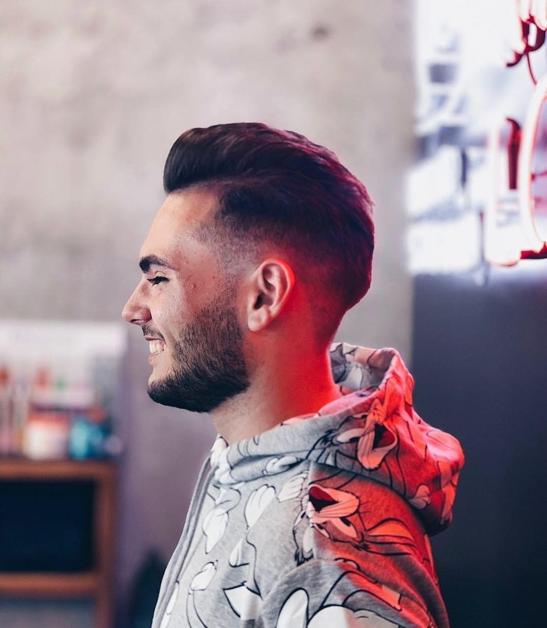 Mężczyzna z modną fryzurą typu fade i krótką brodą, uśmiechający się z profilu, ubrany w bluzę z kapturem w kreskówkowy wzór, na tle betonowej ściany i neonowego oświetlenia.