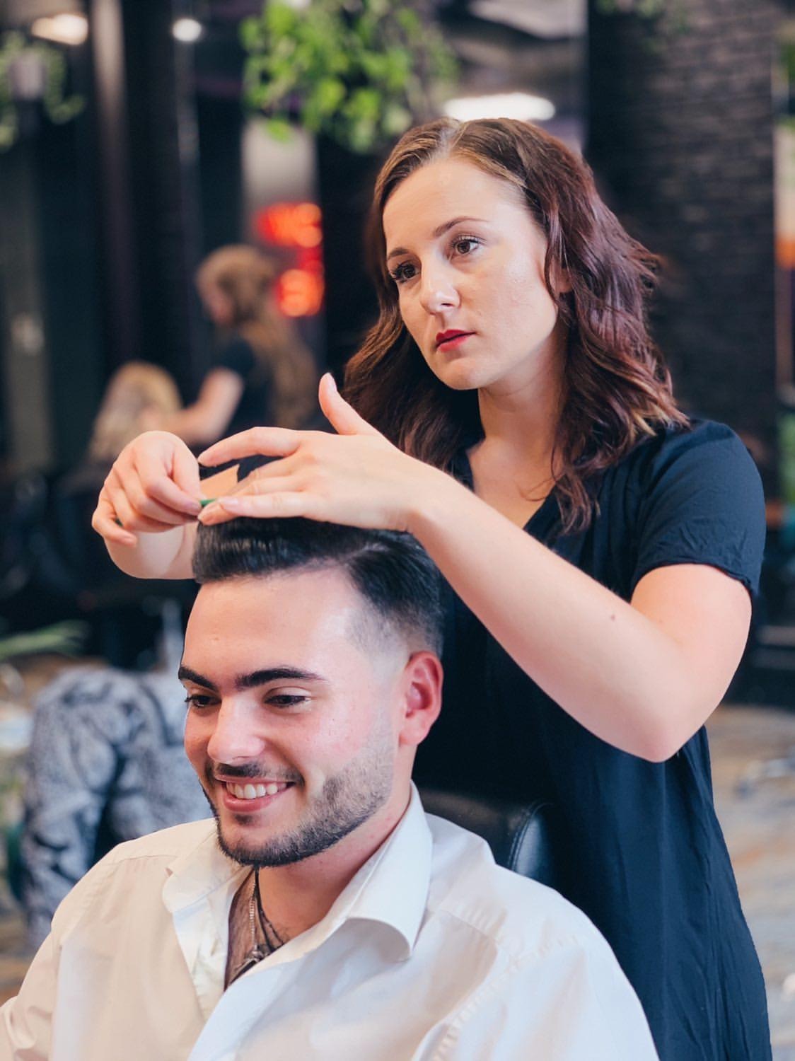 Fryzjerka modeluje fryzurę uśmiechniętemu mężczyźnie w salonie fryzjerskim, używając grzebienia i dłoni do ułożenia włosów.