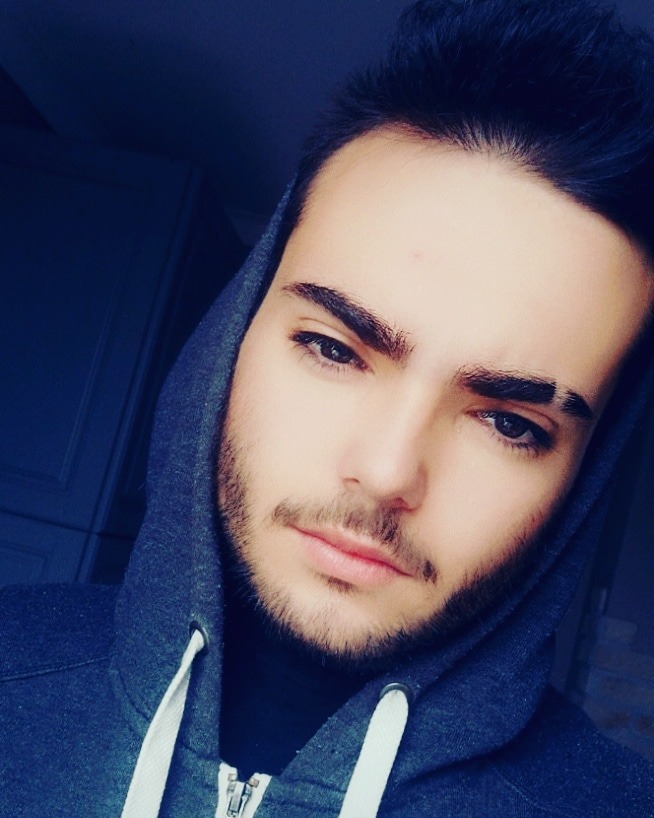 Portret młodego mężczyzny z lekkim zarostem, ubrany w szarą bluzę z kapturem, spoglądającego w dół, z charakterystyczną przerwą w brwi.