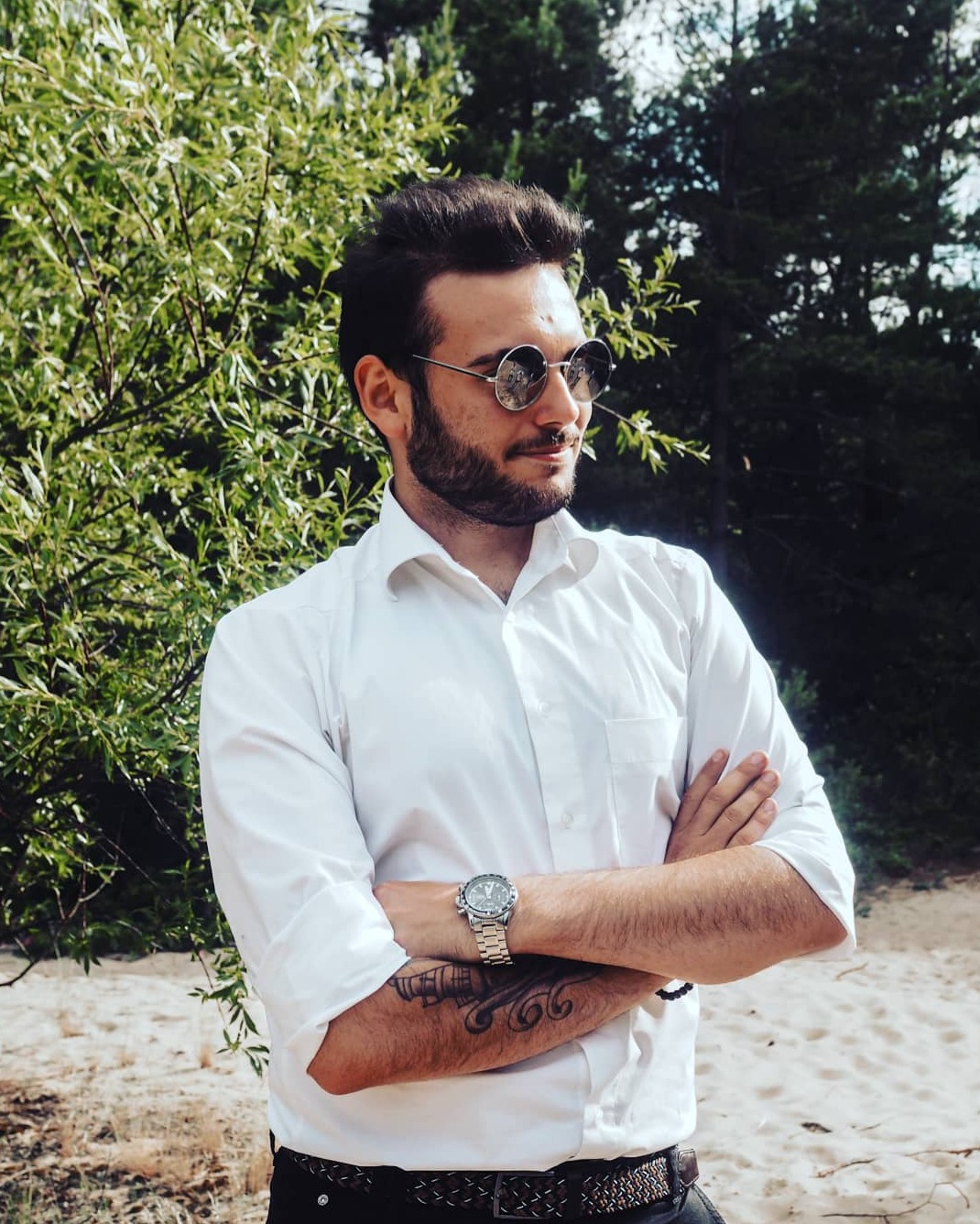 Portret mężczyzny w białej koszuli i okularach przeciwsłonecznych, z tatuażem na ramieniu i zegarkiem, na tle zieleni w Gdańsku.