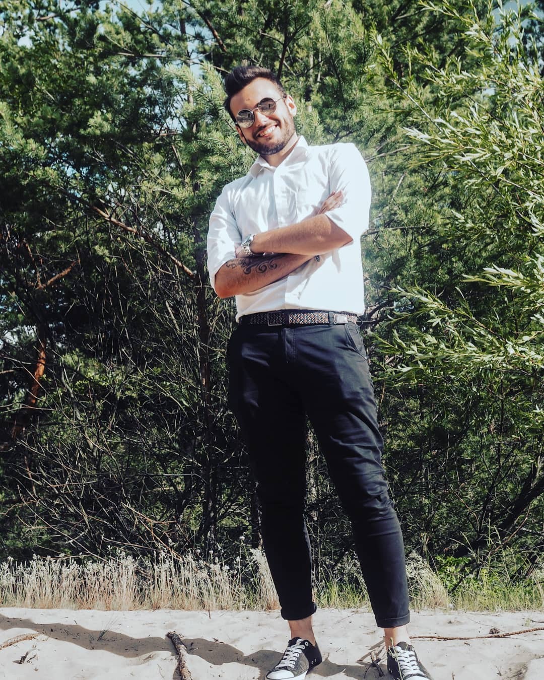 Uśmiechnięty mężczyzna w okularach przeciwsłonecznych, białej koszuli i czarnych spodniach, stoi na piaszczystym podłożu z rękami skrzyżowanymi na tle zieleni drzew.
