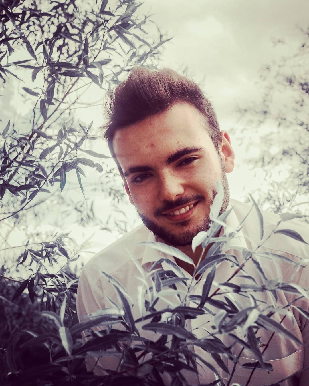 Portret uśmiechniętego mężczyzny w białej koszuli, otoczonego srebrzystymi liśćmi drzewa, z widocznym fragmentem pochmurnego nieba w tle.