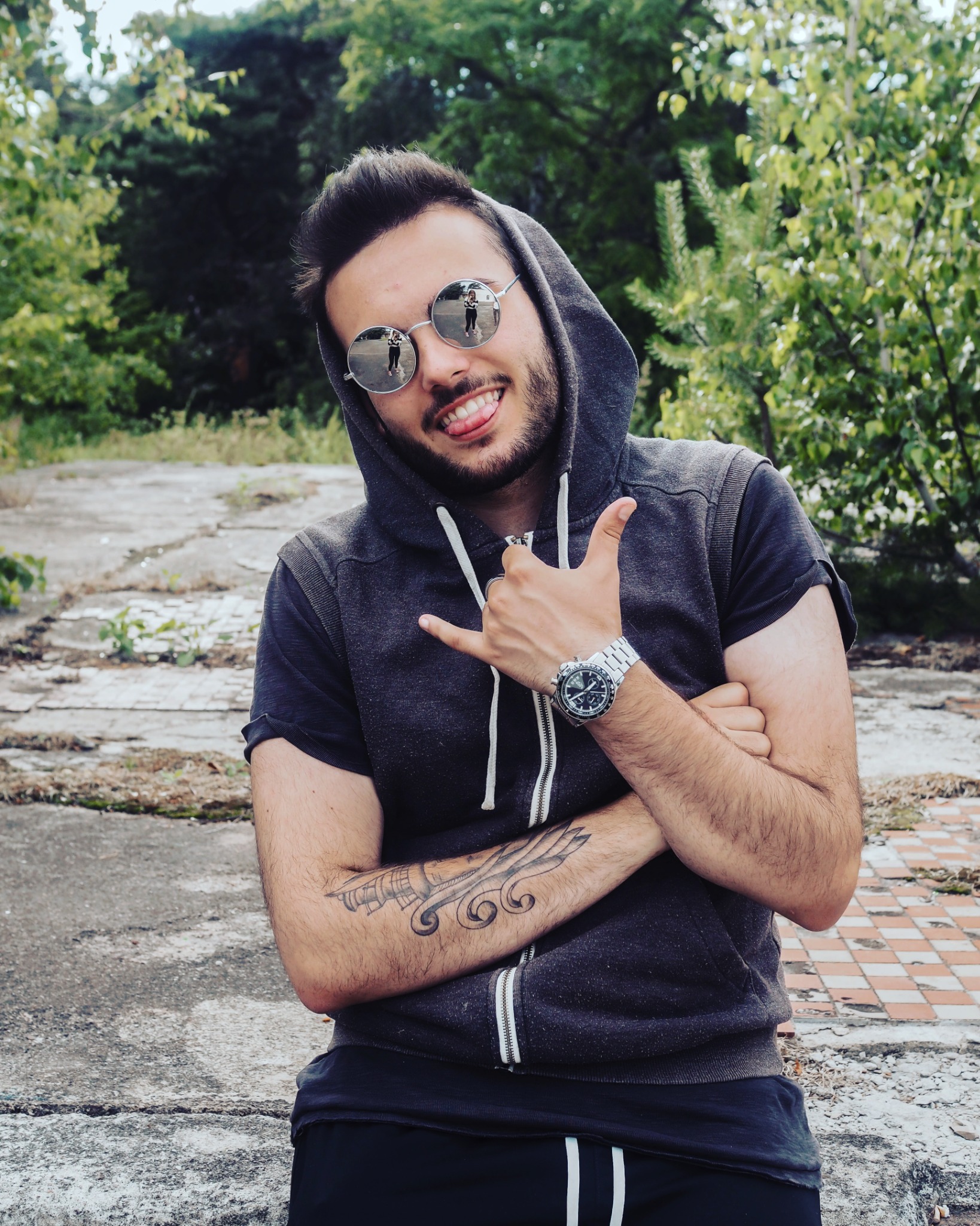 Portret uśmiechniętego mężczyzny w okularach przeciwsłonecznych z odbiciem fotografa, w bluzie z kapturem, z tatuażem na ramieniu, pozującego na tle zieleni i betonowej powierzchni.