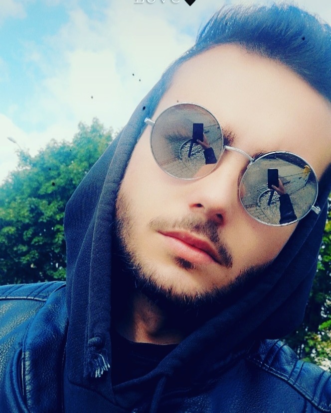 Portret młodego mężczyzny w okularach przeciwsłonecznych z odbiciem fotografa robiącego zdjęcie, ubrany w skórzaną kurtkę i bluzę z kapturem na tle zieleni i nieba.