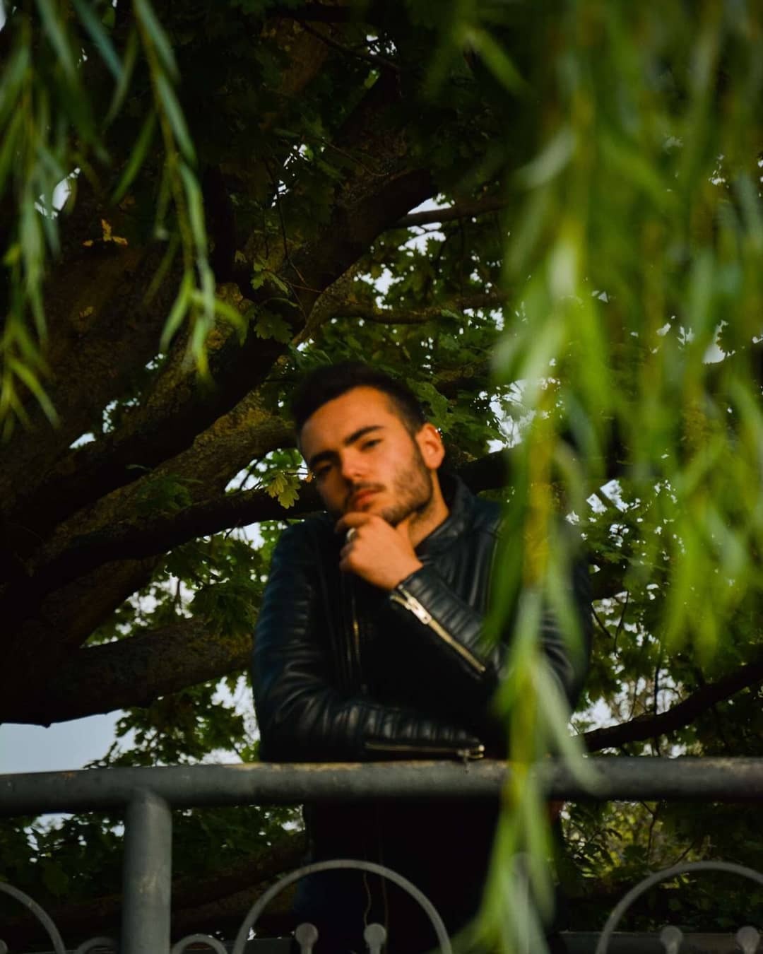 Młody mężczyzna w czarnej skórzanej kurtce oparty o metalową barierkę, zamyślony, z drzewem i zwisającymi gałęziami w tle.