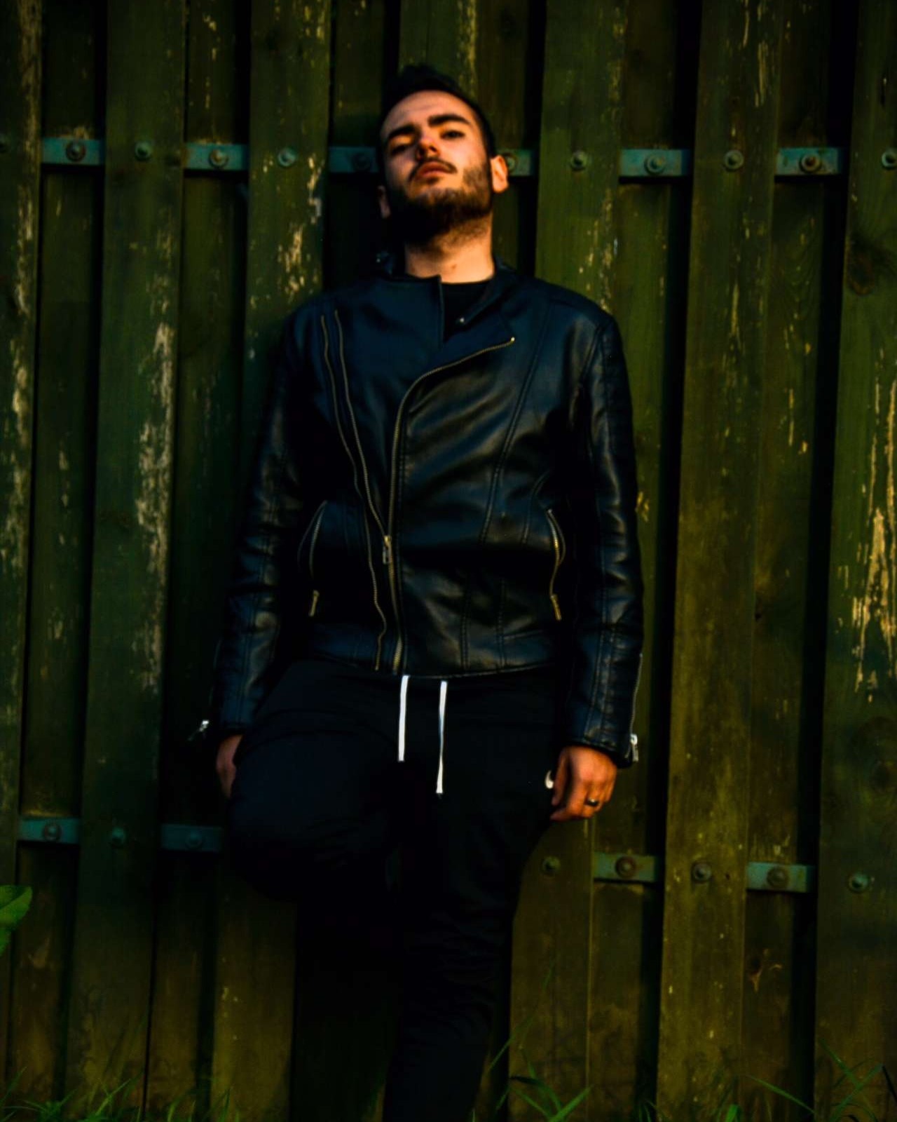 Mężczyzna w czarnej skórzanej kurtce oparty o zielone drewniane ogrodzenie z metalowymi okuciami, ubrany w czarne spodnie dresowe ze sznurkami.