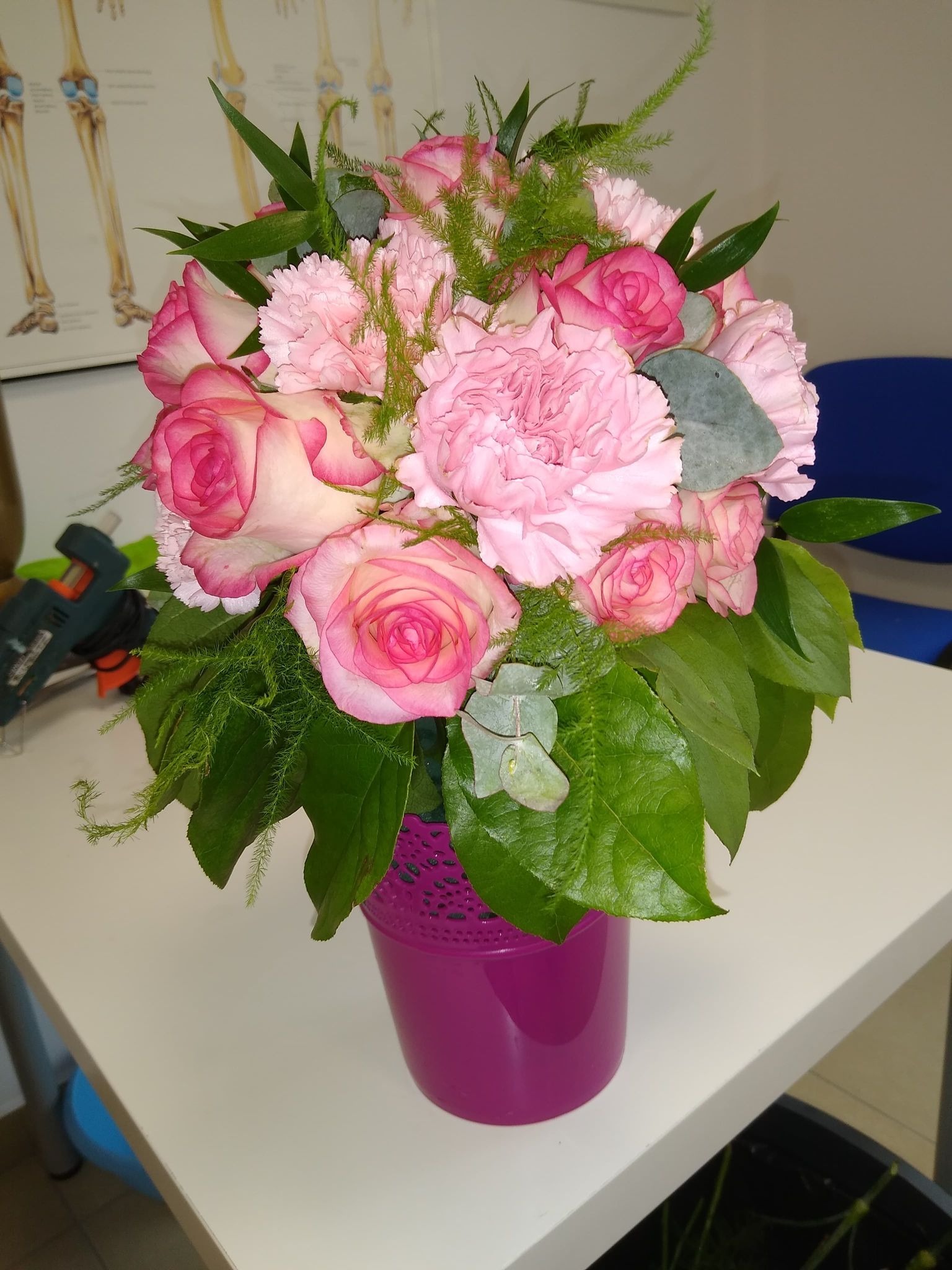 Różowy bukiet z róż i goździków w fioletowym, ażurowym wazonie, widoczny pistolet do kleju i plakat z układem kostnym w tle.