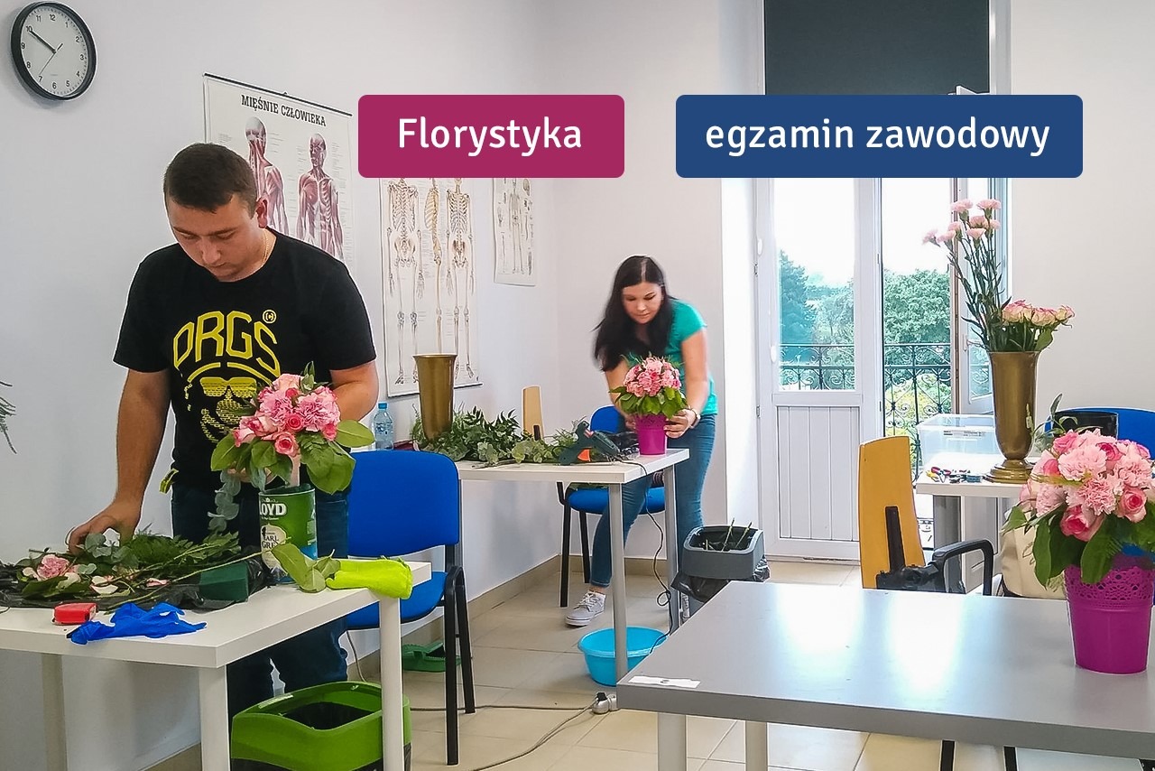 Dwóch florystów, mężczyzna i kobieta, tworzy kompozycje kwiatowe z różowych róż i zielonych dodatków w jasnym pomieszczeniu podczas kursu florystycznego, z widocznym plakatem anatomicznym na ścianie.