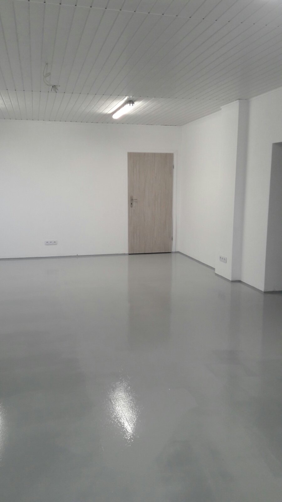 Świeżo wylana, samopoziomująca, szara posadzka żywiczna w pustym pomieszczeniu z białą ścianą, drzwiami i sufitem panelowym z oświetleniem.