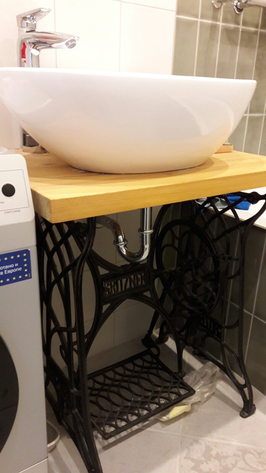 Biała, okrągła umywalka nablatowa na drewnianym blacie, wsparta na odrestaurowanej, żeliwnej podstawie maszyny do szycia marki Britzner, z widoczną chromowaną armaturą i fragmentem pralki obok.