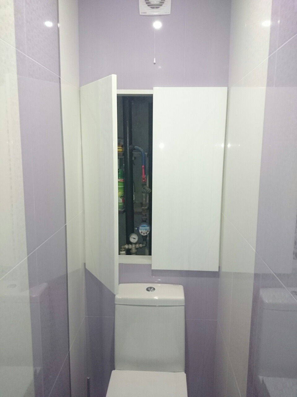 Toaleta w łazience z fioletowymi płytkami, nad nią szafka z ukrytymi rurami i licznikami.
